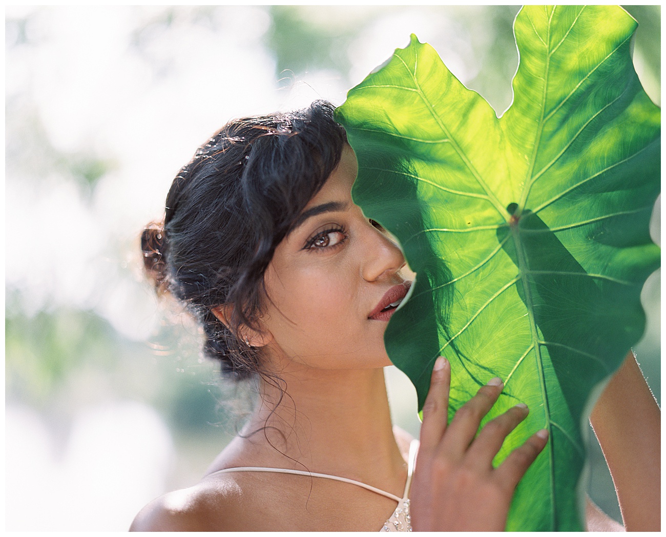 Gorgeous Indian Bride with large tropical leaves and greenery basking in the backlight of the sunlight for her wedding day wearing tadashi bridal gown.