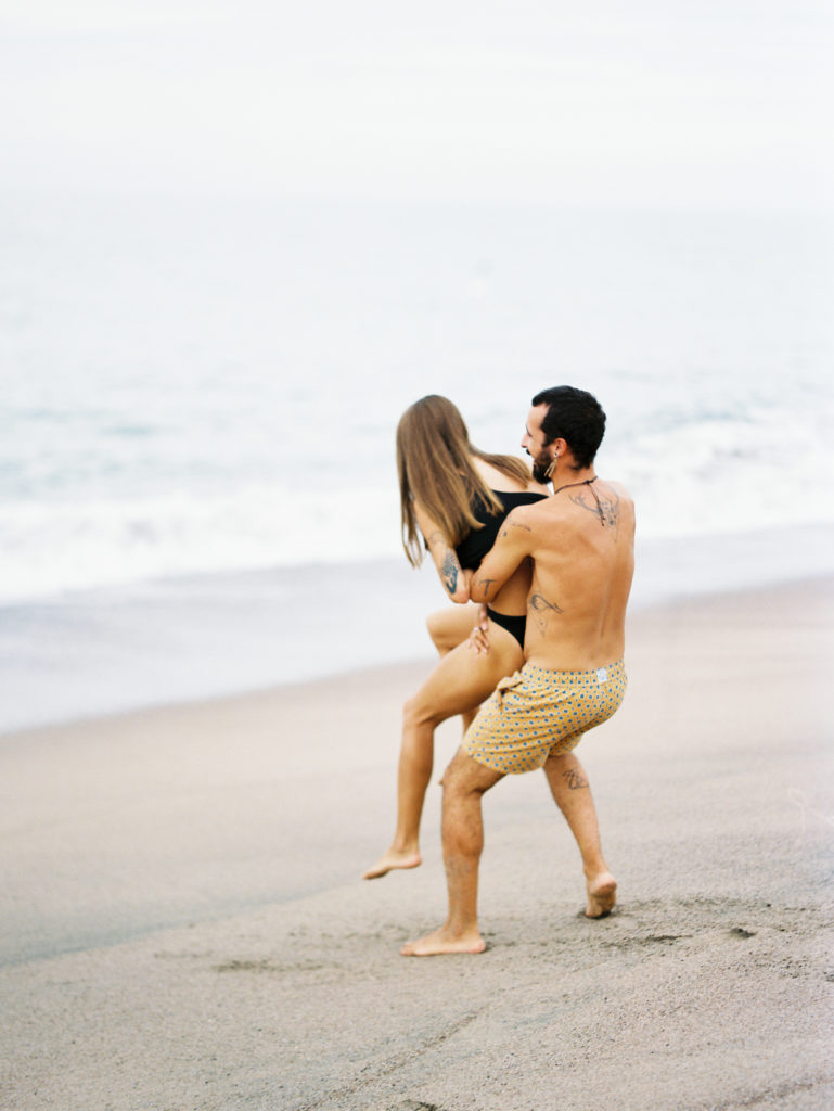 laughing couple on a beach in mexico, playing and throwing eachother into the water, wearing neutral swim wear of black and gold.