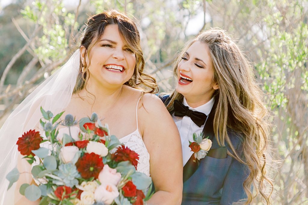 Laughing LGBTQ+ brides together on their wedding day with red and white florals and a blue custom suit.